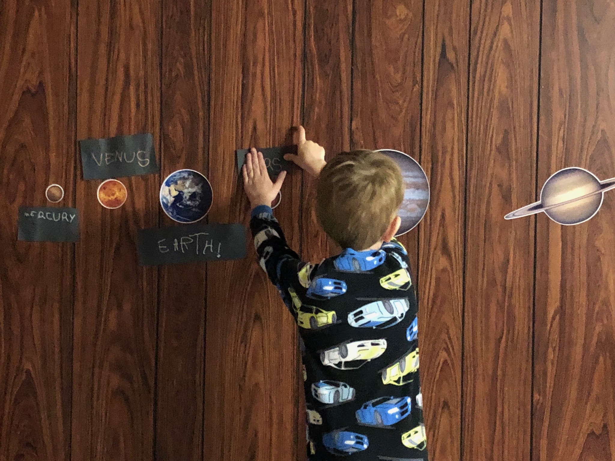 A young boy is sticking a black label for Mars up on a wall. Planet wall stickers and labels can be seen for Mercury, Venus and Earth