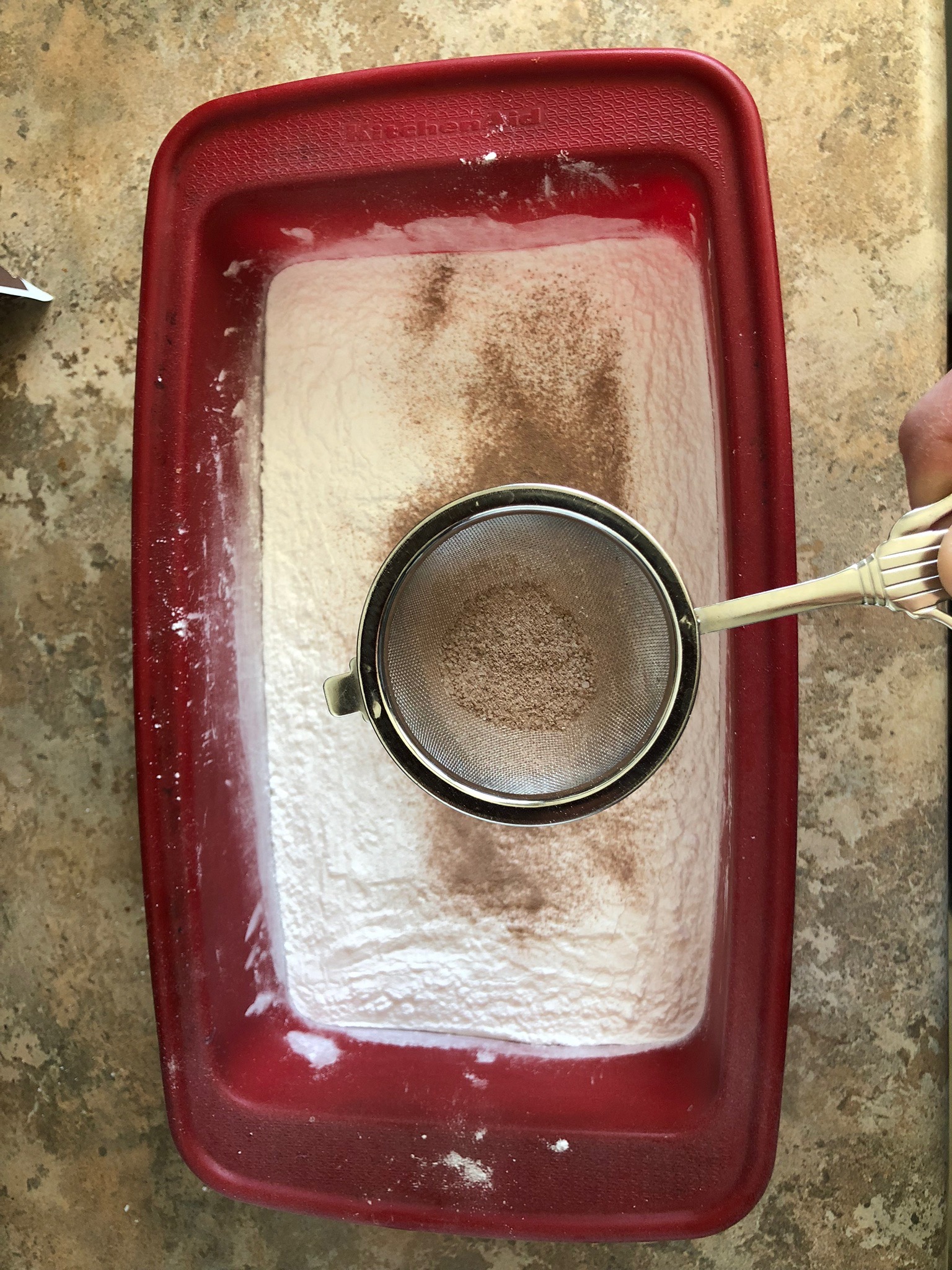 A strainer taps cocoa into a silicone bread pan filled with flour,