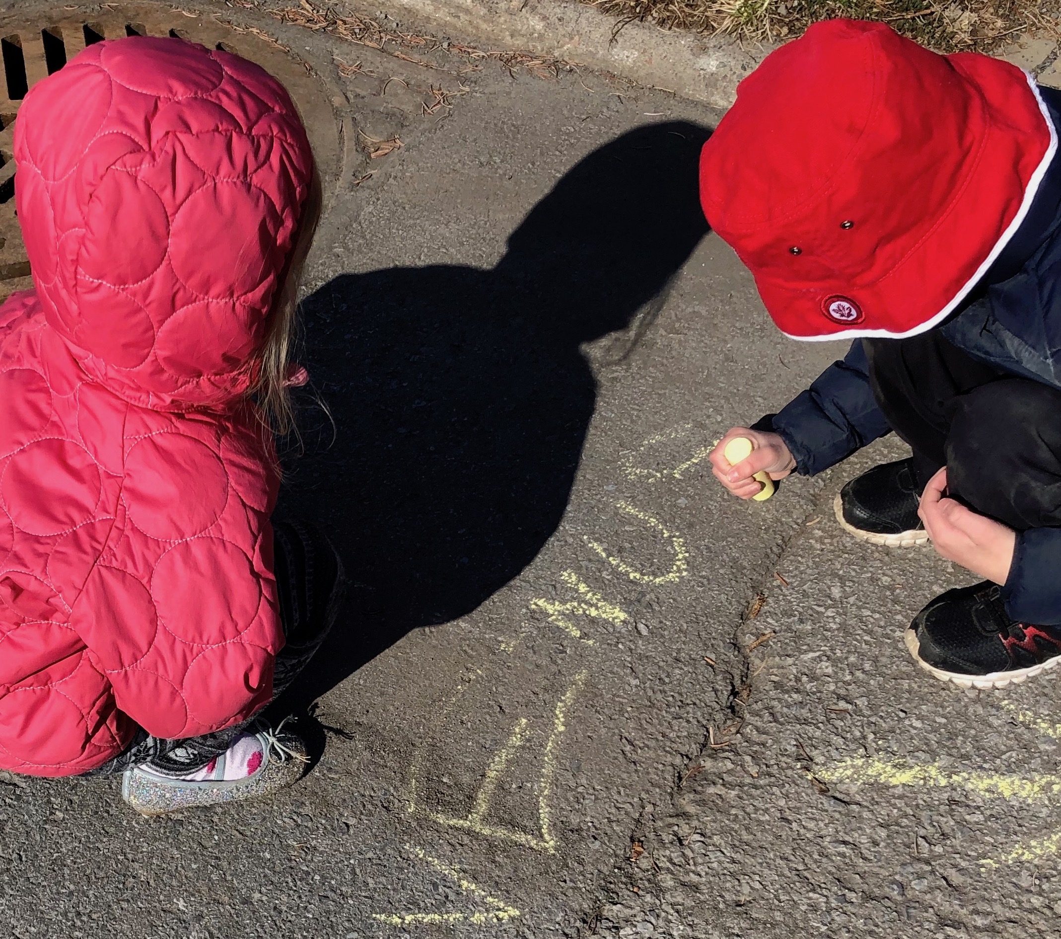 A young girl and a young boy look at the ground as the boy writes Venus in chalk. The girl's shadow is seen above Venus