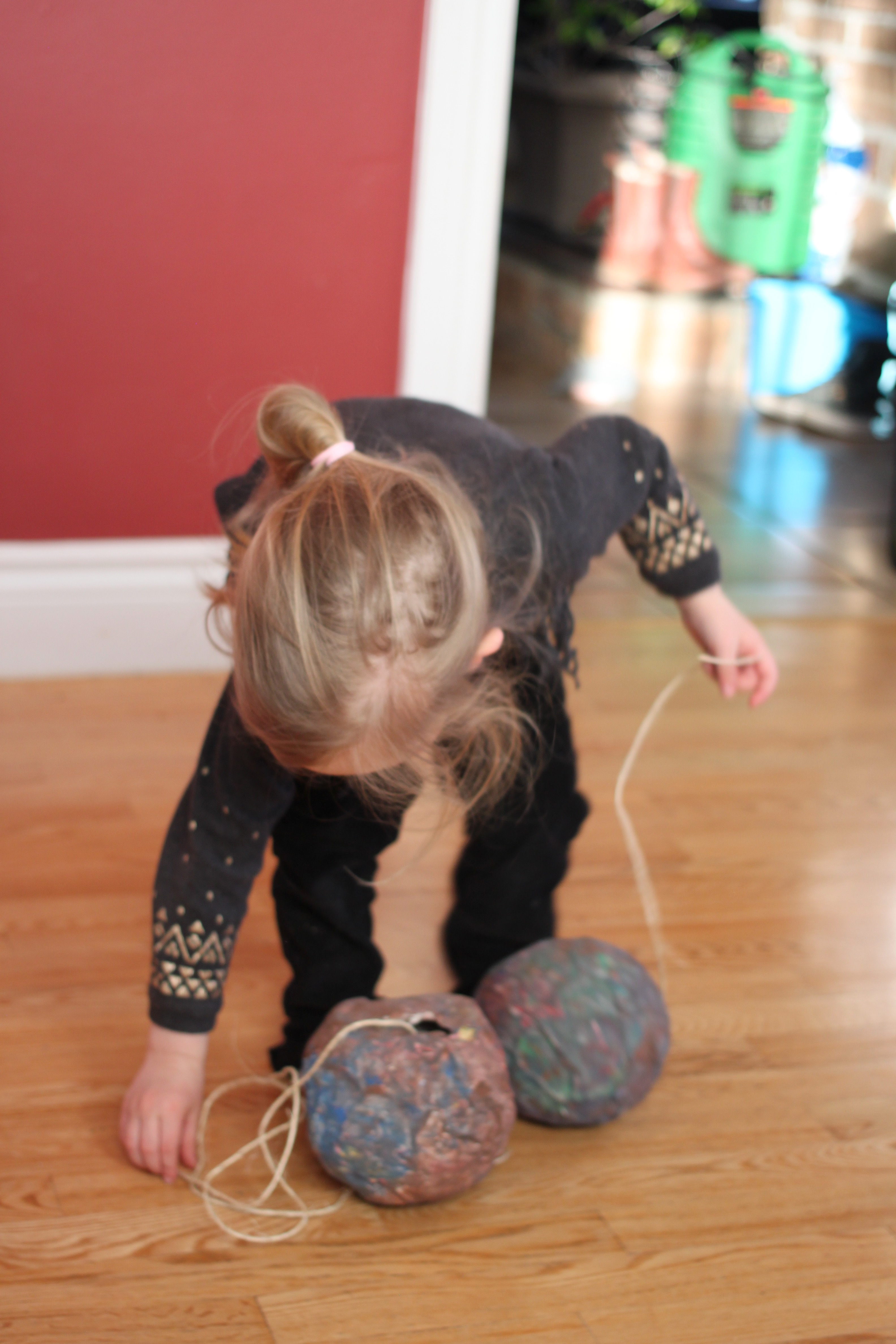 A young girl is picking up a string from the ground, attached to a paper mâché asteroid. She holds another string with another asteroid in her other hand