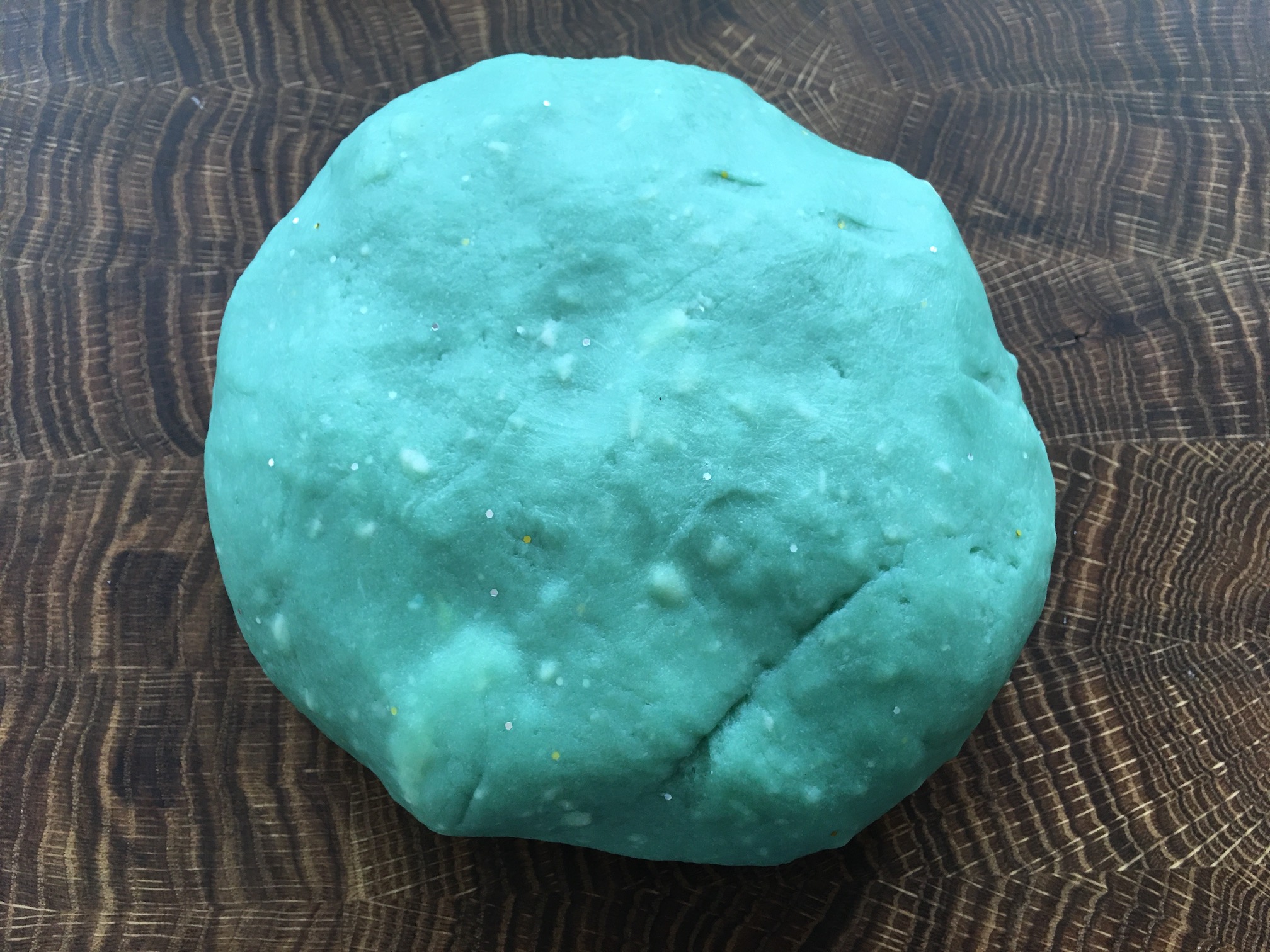 A lumpy turquoise blob of turquoise glitter play dough sits on a wooden board.