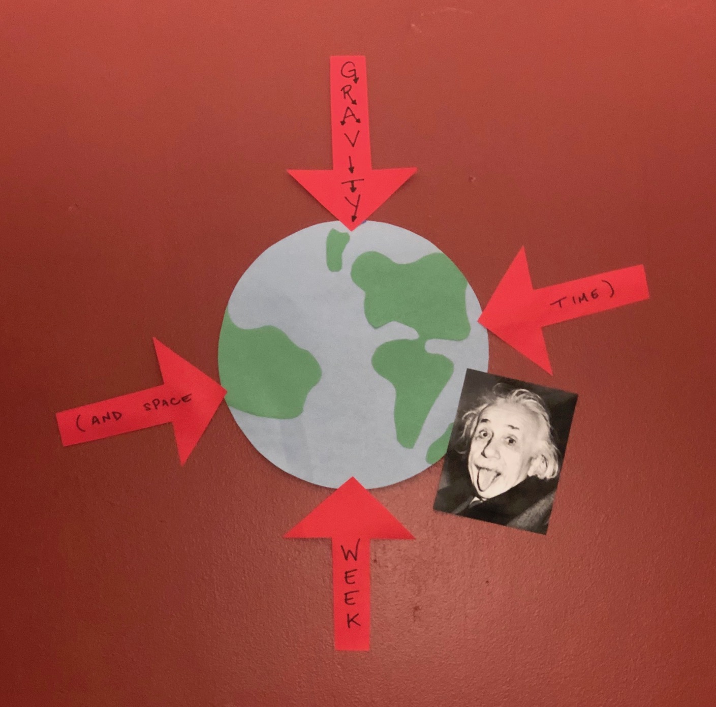 An earth made of construction paper is pasted on a wall. Four red arrows point towards the earth, each with part of the sentence Gravity and Spacetime Week. A picture of Albert Einstein with his tongue out overlaps part of the earth. They are all based on a dark red wall.