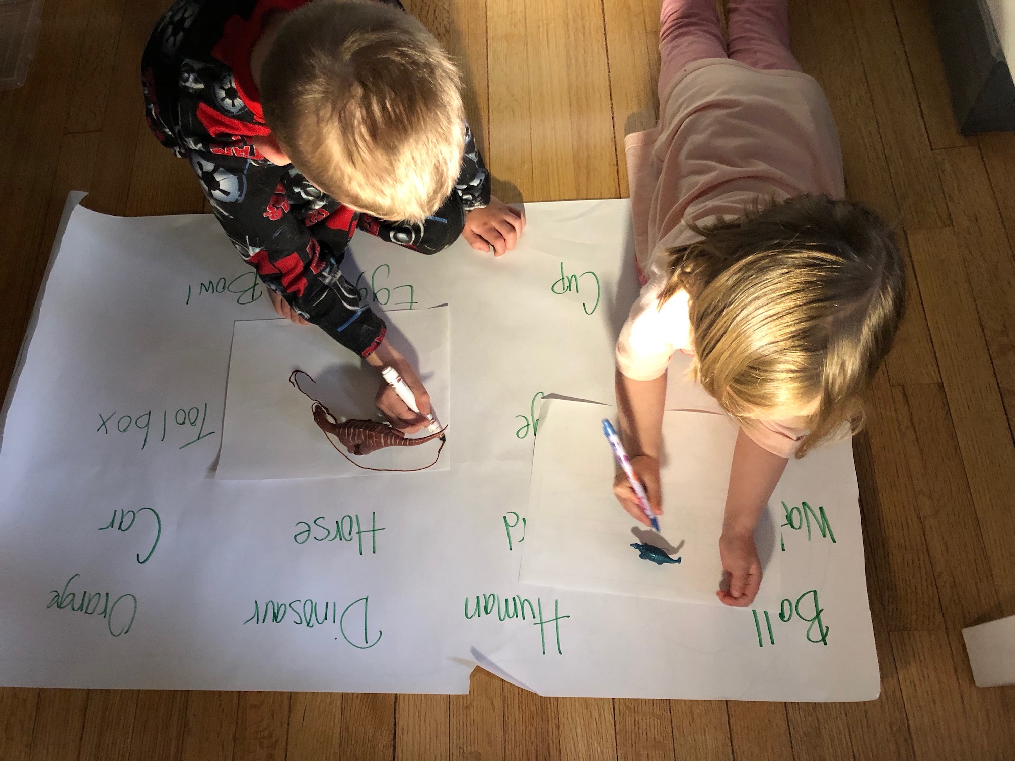 Two young children are seen drawing on white pieces of paper. Each paper has a plastic dinosaur, with a light casting a shadow on the paper.