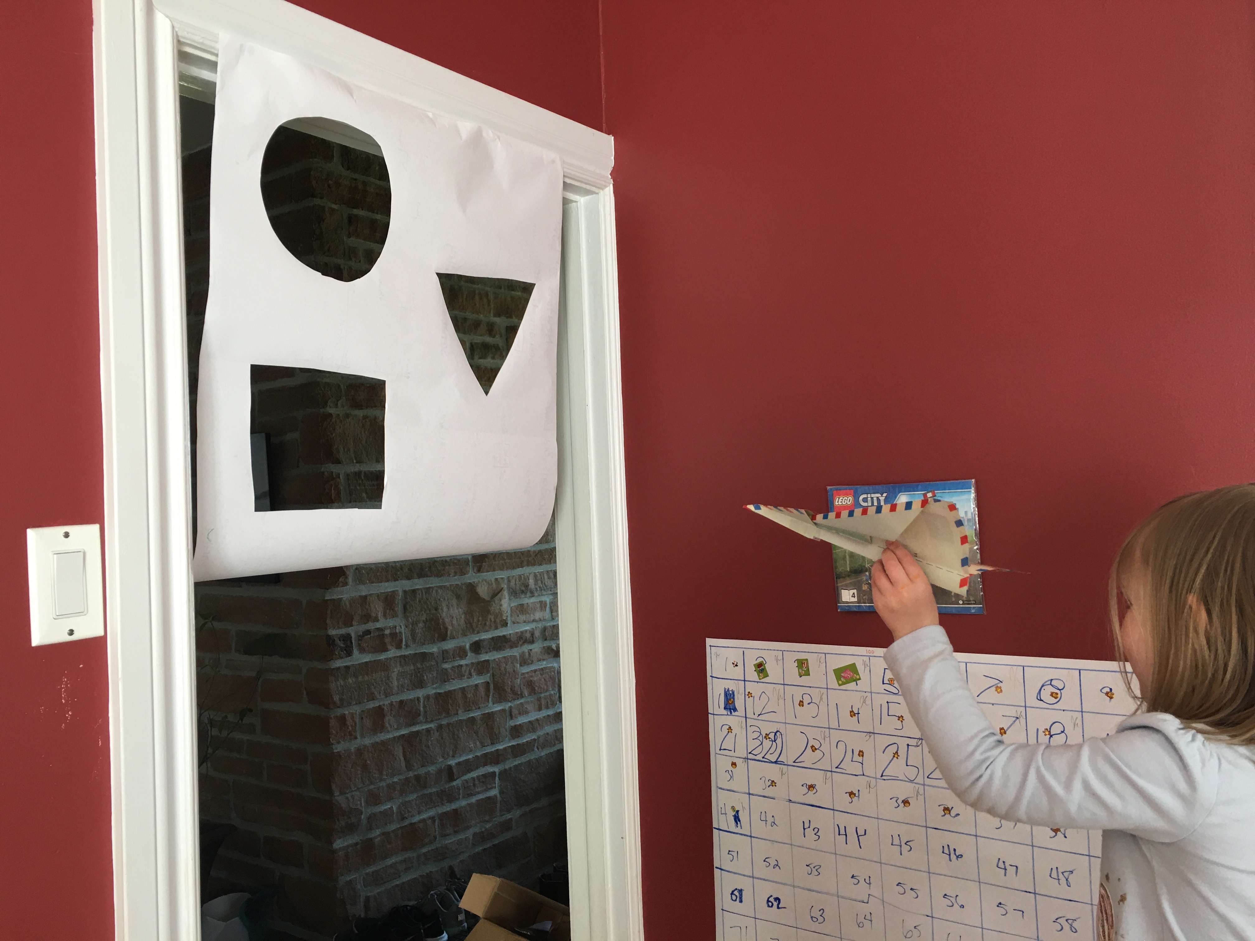 A little girl is seen holding a paper airplane ready to throw it towards a large white paper hanging from a doorframe, with a circle, triangle, and rectangle cut out as targets