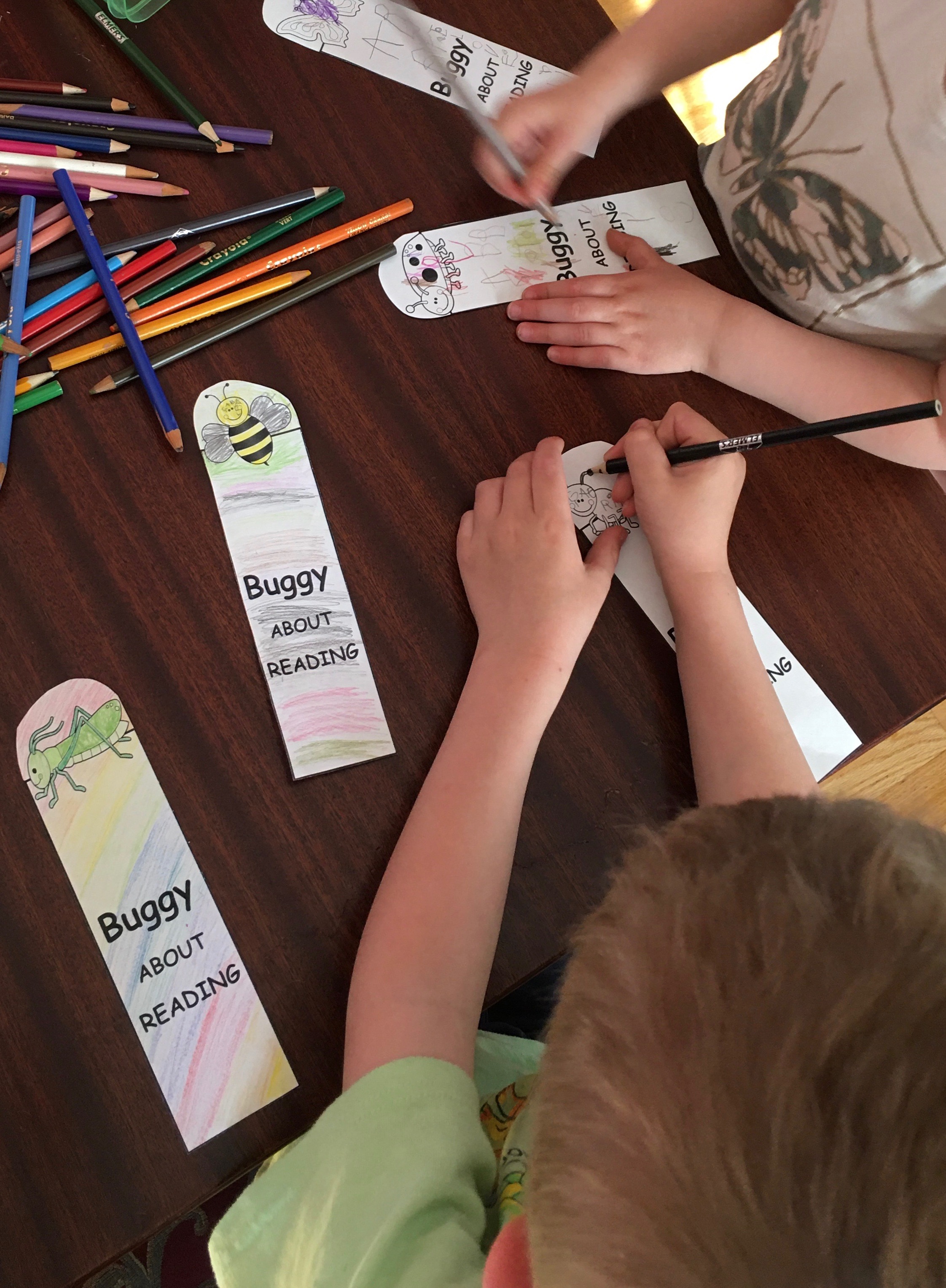 Two children are seeing colouring in paper bookmarks that read Buggy About Reading. Each bookmark has an insect on it.