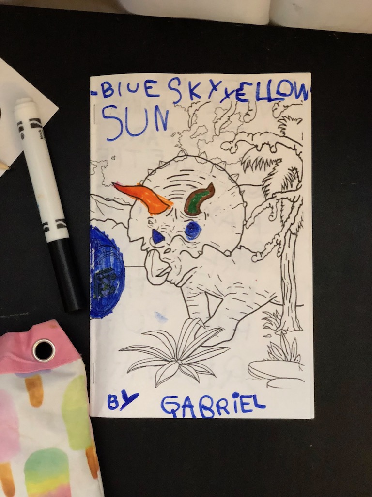 A triceratops printed in black and white is partially coloured in. The text Blue Sky Yellow Sun by Gabriel has been printed by a young child.